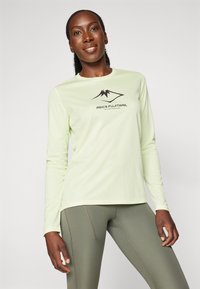 Hymyilevä nainen, joka pitää päällään vaaleanvihreää pitkähihasta Asics Fujitrail-paitaa ja oliivinvihreitä legginssejä, seisoo valkoista taustaa vasten.