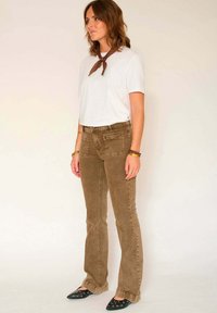 Pantalon évasé en velours côtelé marron avec deux poches avant et arrière, associé à une chemise blanche à manches courtes et un foulard marron autour du cou. Chaussures plates noires à bout pointu.