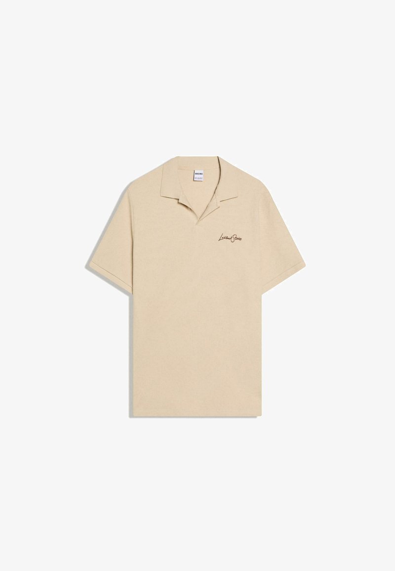 Polo beige à manches courtes avec col rabattable et petit logo en texte cursif noir sur le côté gauche de la poitrine.
