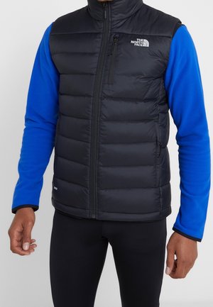 Mężczyzna ubrany w czarną pikowaną kamizelkę bez rękawów z kieszeniami na zamek, na niebieskiej koszuli z długim rękawem i czarnych spodniach, stojący z opuszczonymi rękami.