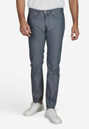 Man die een nauwsluitende grijze broek, witte sneakers en een wit overhemd draagt, staand tegen een effen witte achtergrond.