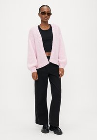 Γυναίκα που στέκεται φορώντας γυαλιά ηλίου με σχέδιο χελώνας, ροζ ανοιχτό πλεκτή ζακέτα, μαύρο κοντό τοπ, μαύρο φαρδύ παντελόνι και μαύρα παπούτσια με ψηλή σόλα.