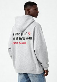 Pelēks džemperis ar kapuci, ar melnu un sarkanu tekstu aizmugurē. Izstrādāts brīvai piegādei, ar ribotām piedurknēm un apakšmalu.