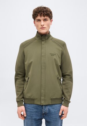 Junger Mann mit kurzem lockigem Haar, der eine olivgrüne Jacke mit Reißverschluss und blaue Jeans trägt, vor einem schlichten hellen Hintergrund stehend.