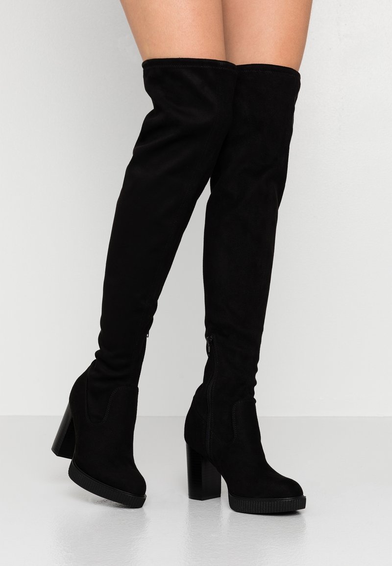 Bottes noires au-dessus du genou en suède, avec un talon haut épais, une fermeture éclair latérale et une texture lisse.