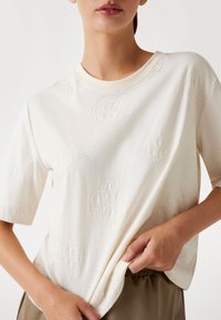 Korte mouwen crème katoenen t-shirt met reliëf rond en bloemig patroon, wijde halslijn en een losse pasvorm, gecombineerd met olijfgroene broek.