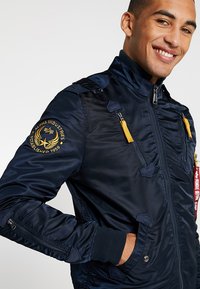 Navy bomber jacket with a zip closure, ribbed cuffs, and multiple zippered pockets. Features an embroidered logo and yellow zip pulls.