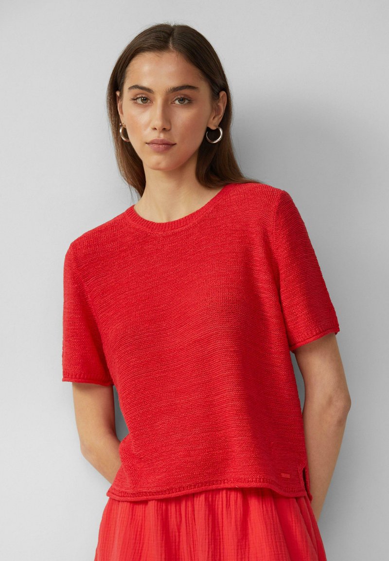 Rode gebreide top met korte mouwen en een ronde halslijn, voorzien van een gestructureerde afwerking en een iets losse pasvorm. De onderkant heeft een kleine zijsplit.