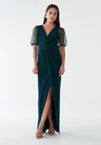 Tumši zaļa kleita ar savītu priekšpusi, caurspīdīgām pūkām un augsto šķēlumu. Gluda auduma forma ar smalku tekstūru un spīdumu.