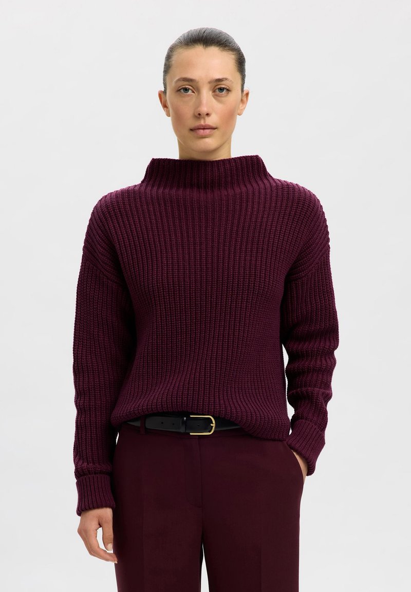Pull en maille bordeaux avec un col haut, texture côtelée et coupe décontractée, associé à un pantalon assorti et une ceinture noire avec une boucle dorée.