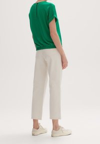 Grøn, kortærmet top med elastisk kant og knapdetalje på ryggen; parret med hvide cropped bukser og lyse sneakers.