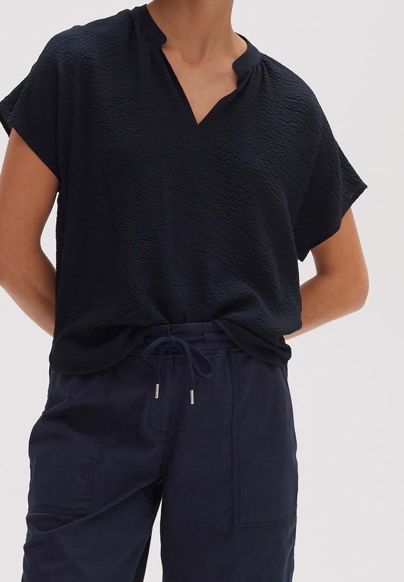 Persoon in een zwarte top met korte mouwen en V-hals van textuur, met marineblauwe broek met trekkoord en voorzakken, staand met de handen achter de rug.