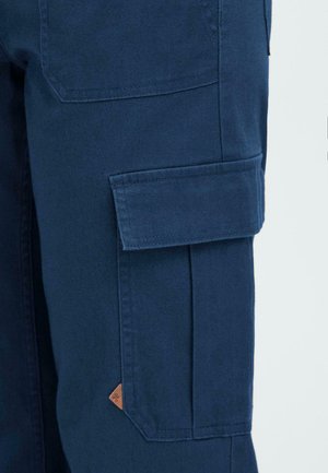 Dark blue cargo pants close-up showing side flap pocket with stitching and small brown triangular logo tab.