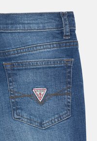 Jean en denim bleu avec un patch triangulaire rouge portant le logo "GUESS" sur la poche arrière, avec des coutures visibles et un design classique à cinq poches.