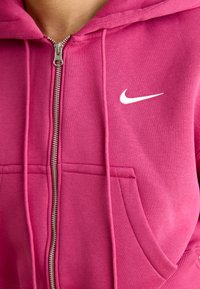Ružový zipsový mikina s strieborným zipsom, prednými vreckami a bielym logom Nike umiestneným na ľavej hrudnej oblasti. Mäkko tkaninová textura.