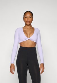 Fialový dlhý rukáv crop top s hlbokým V-výstrihom a prekrúteným predným dizajnom, v kombinácii s čiernymi legínami s vysokým pásom. Hladká textúra látky.