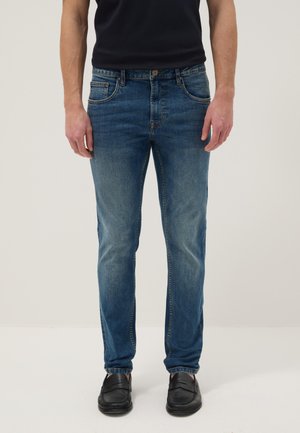 Man draagt mediumblauwe slim-fit jeans, zwarte loafers en een zwart overhemd, staand tegen een effen lichte achtergrond.