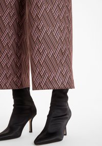 Un pantalon large à motifs présente un design géométrique rayé en rose et marron. Des bottes à talons aiguilles pointues noires avec une texture douce et un talon fin.