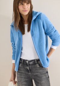 Femme portant une veste polaire zippée bleu clair sur un t-shirt blanc et un jean gris, tenant un sac blanc, debout contre un mur clair.