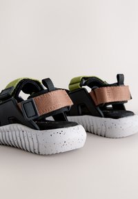 Sandales noires et grises avec des sangles réglables vertes et marron, dotées d'une semelle blanche tachetée texturée et d'un design durable.