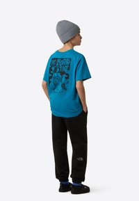 Turkizna majica s krepko črno grafično zasnovo na hrbtu, združena s črnimi trenirkami in temnimi supergami. Siva kapa na glavi.