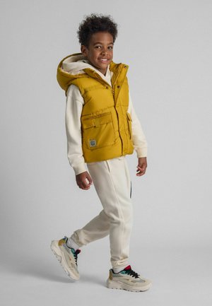 Gilet en duvet jaune avec une capuche, comportant deux poches avant et un patch de logo. Porté sur un sweatshirt blanc et un pantalon assorti, avec des baskets colorées.
