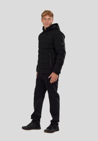 Chaqueta de plumas negra con capucha, con costuras horizontales, bolsillos con cremallera y un diseño elegante. Llevada sobre jeans negros y zapatos.