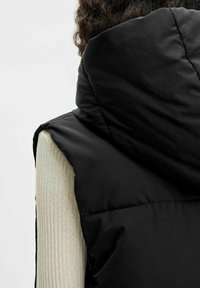 Veste noire matelassée avec un col montant et une capuche. Elle présente une texture lisse et des ouvertures latérales, portée par-dessus un haut côtelé clair.