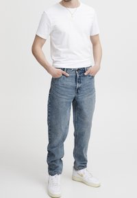 Homme portant un t-shirt blanc, un jean bleu, des baskets blanches avec un swoosh doré, et un collier pendentif doré, debout les mains dans les poches.