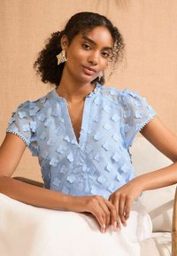 Blusa azzurra chiara a maniche corte con applicazioni floreali testurizzate, chiusura a bottoni e cuciture intricate. Tessuto liscio con una leggera lucentezza.