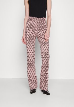 Femme portant un pantalon taille haute à jambes droites avec un motif géométrique rose et noir, associé à des chaussures pointues noires et un haut noir sans manches.