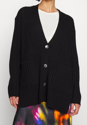 Femme portant un cardigan côtelé noir avec de gros boutons par-dessus un top en maille blanche et une jupe abstraite colorée.