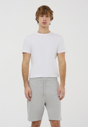 Maglietta bianca a girocollo abbinata a pantaloni corti grigi chiari con cordino, entrambi realizzati in tessuto morbido, caratterizzati da una vestibilità comoda e pantaloni corti fino al ginocchio.