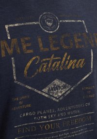 Navy blue cotton t-shirt featuring metallic gold and white text, with graphic elements and a vintage aviation theme, including shield shapes.