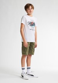 Camiseta blanca con un estampado gráfico en azul y naranja, combinada con pantalones cortos de cargo caqui, zapatillas blancas con detalles en negro y calcetines de rayas.