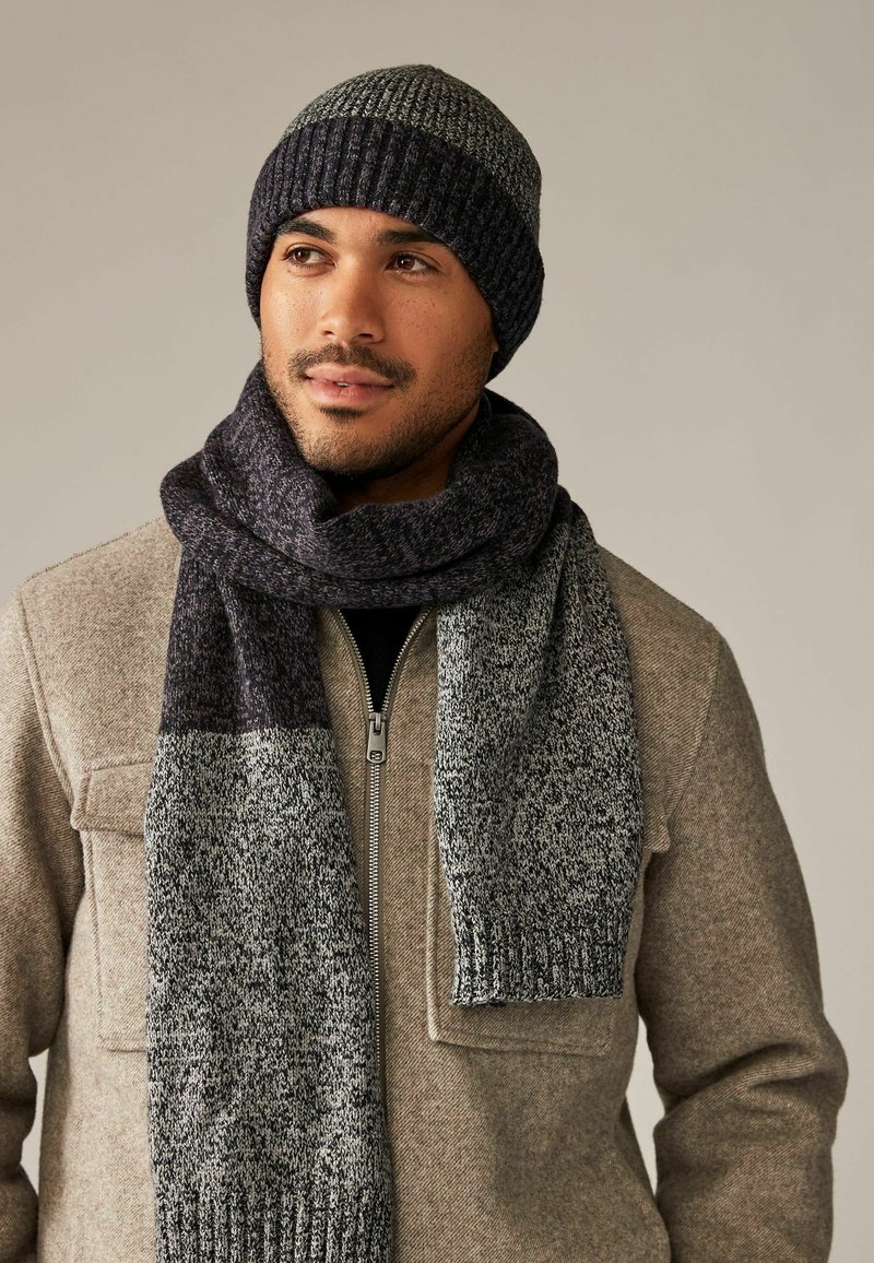 Virkat mössa och scarf-set i mörkgrått med ribbat mönster. Scarfen har en tvåfärgad design och är kombinerad med en beige jacka.
