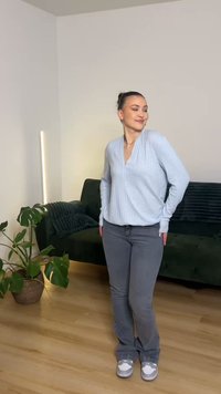 Light blue ribbed long-sleeve top with a v-neck, paired with grey flared jeans and grey sneakers. Indoor setting with a green sofa and plants.
