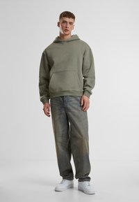 Sudadera de algodón verde con bolsillo canguro, combinada con jeans azules desgastados de corte holgado y zapatillas blancas. Diseño sencillo y casual.