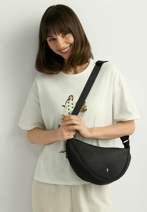 Młoda kobieta o brązowych włosach do ramion uśmiecha się, ubrana w białą koszulkę z grafiką i beżowe spodnie, trzyma czarną torebkę na ramię na tle białej ściany.