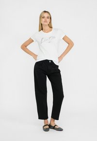 Hvid t-shirt med sølv "Luca" tekst, sorte loose-fit jeans med perleudsmykning på lommen og sorte flade sko. Enkel, afslappet outfit.