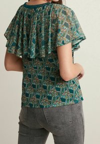 Grüne, gemusterte Bluse mit Rüschensleeves, die florale und geometrische Designs aufweist, kombiniert mit grauen Jeans. Textil mit einer dezenten glänzenden Oberfläche.