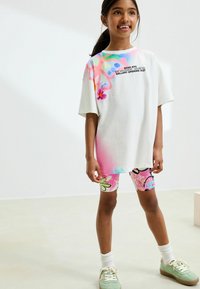 T-shirt oversize blanche avec des éclaboussures roses et bleues et du texte noir, associée à un short de vélo coloré à motifs et des baskets vertes.