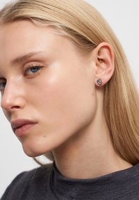 Orecchini d'argento a forma di cuore, con un design elegante. La modella ha i capelli biondi dritti di lunghezza media e indossa un top grigio, vista di profilo.