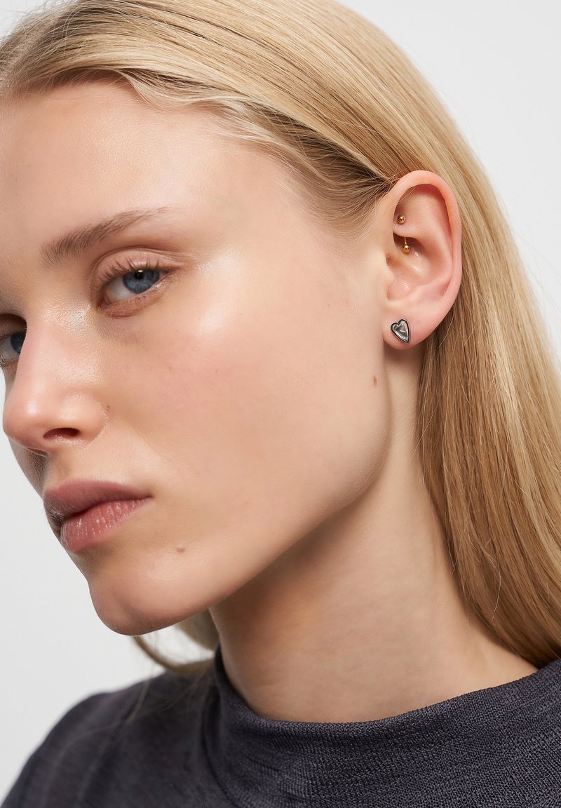 Orecchini d'argento a forma di cuore, con un design elegante. La modella ha i capelli biondi dritti di lunghezza media e indossa un top grigio, vista di profilo.