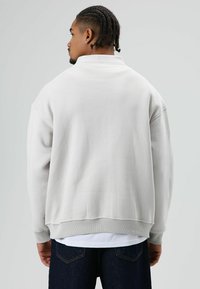 Lichtgrijze pullover met hoge hals, doorgezakte schouders en geribbelde manchetten. Gladde textuur en een ontspannen pasvorm, gedragen over donkere jeans.