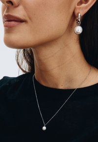 Sudraba rotaslietas ar apaļo pērļu piekari uz plānas ķēdītes un pērļu dekoriem rotātu auskaru gredzenu, uz melnas kreklas fona.