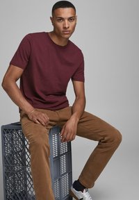 Burgunderfarbene Kurzarm-T-Shirt kombiniert mit braunen Hosen. Das Model sitzt auf einer grauen Kunststoffkiste und trägt weiße Sneaker mit blauen und roten Akzenten.