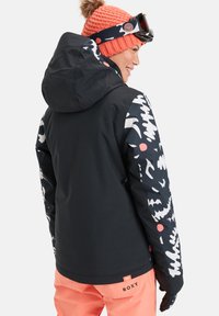 Casaco preto com mangas de tecido estampado, apresentando designs ousados em branco e coral. Combinado com calças corais e um gorro de malha laranja.