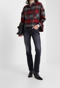 Camisa de cuadros roja y gris con dos bolsillos en el pecho, usada sobre un suéter de cuello alto beige. Combinada con jeans oscuros y botas de tacón negro hasta el tobillo.