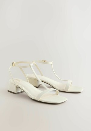 Białe skórzane sandały na słupku z paskiem T, kwadratowym noskiem i teksturowanym detalem na obcasie o gładkim wykończeniu. Złote akcenty hardware.